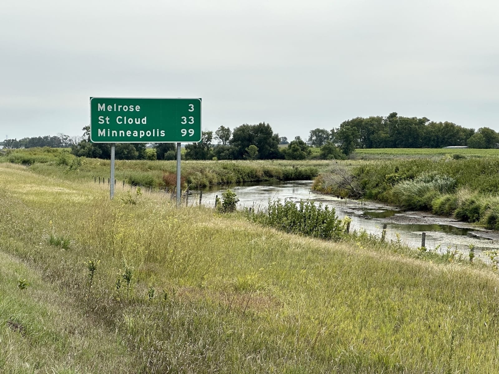 A green distance sign by a creek: Melrose 3, St Cloud 33, Minneapolis 99.