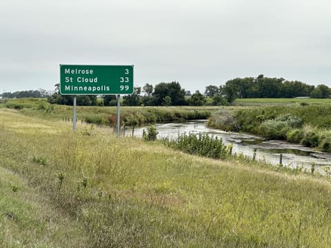 A green distance sign by a creek: Melrose 3, St Cloud 33, Minneapolis 99.