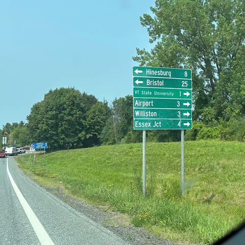 A green guide sign beside the road: Hinesburg 8, Bristol 25, VT State University 1, Airport 3, Williston 3, Essex Jct 4.